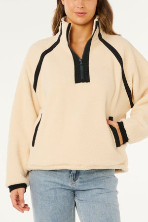 Wearing the Rip Curl Surf Club Heritage Polar fleece pullover in cream with black trim and a half-zip front, paired with light blue jeans, a person poses with one hand in their pocket against a white background.