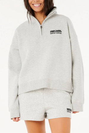 A woman smiles in a light gray Rip Curl Surf Puff 1/4 Zip sweatshirt and matching shorts with "Ripcurl Surfing Company" logos, creating a casual and comfortable look ideal for outdoor adventures.