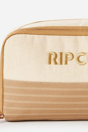Close-up of a beige and light brown Rip Curl Surf Revival Toiletry Bag with the brand name "RIPCURL" embroidered in gold lettering on the front. The top half of this recycled polyester toiletry bag is solid beige, while the bottom half features a striped light brown pattern.