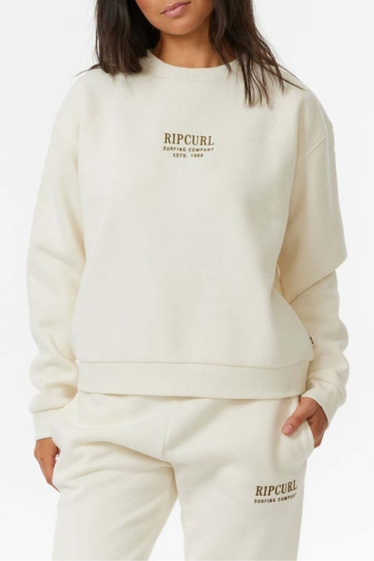 A woman stands against a plain white background wearing an off-white Rip Curl Surf Staple Relaxed Crew sweatshirt and matching fleece-lined sweatpants, both featuring the brand logo and "Rip Curl Surfing Company Estd. 1969.
