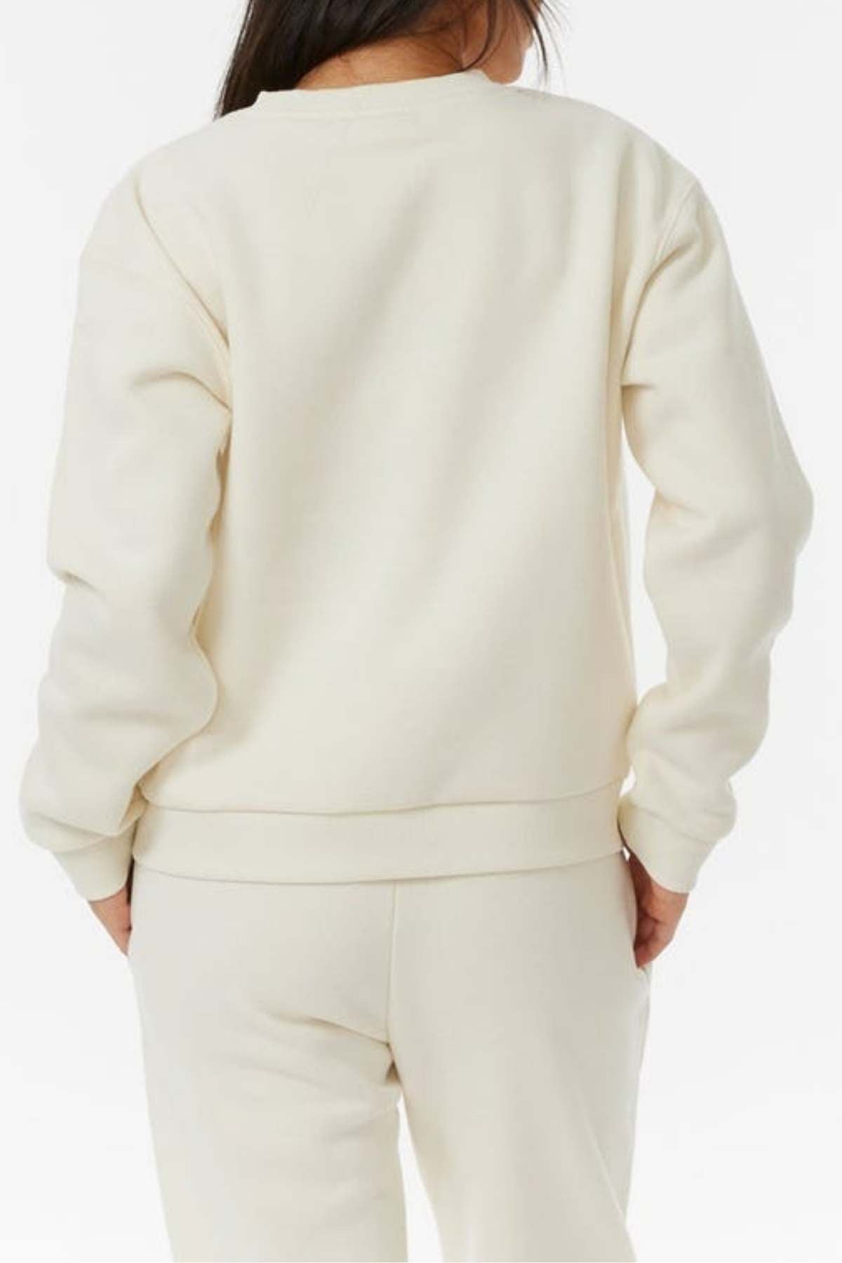 A person with long dark hair is shown from the back wearing a Rip Curl Surf Staple Relaxed Crew sweatshirt and matching sweatpants in off-white, both in a relaxed fit, against a plain white background.