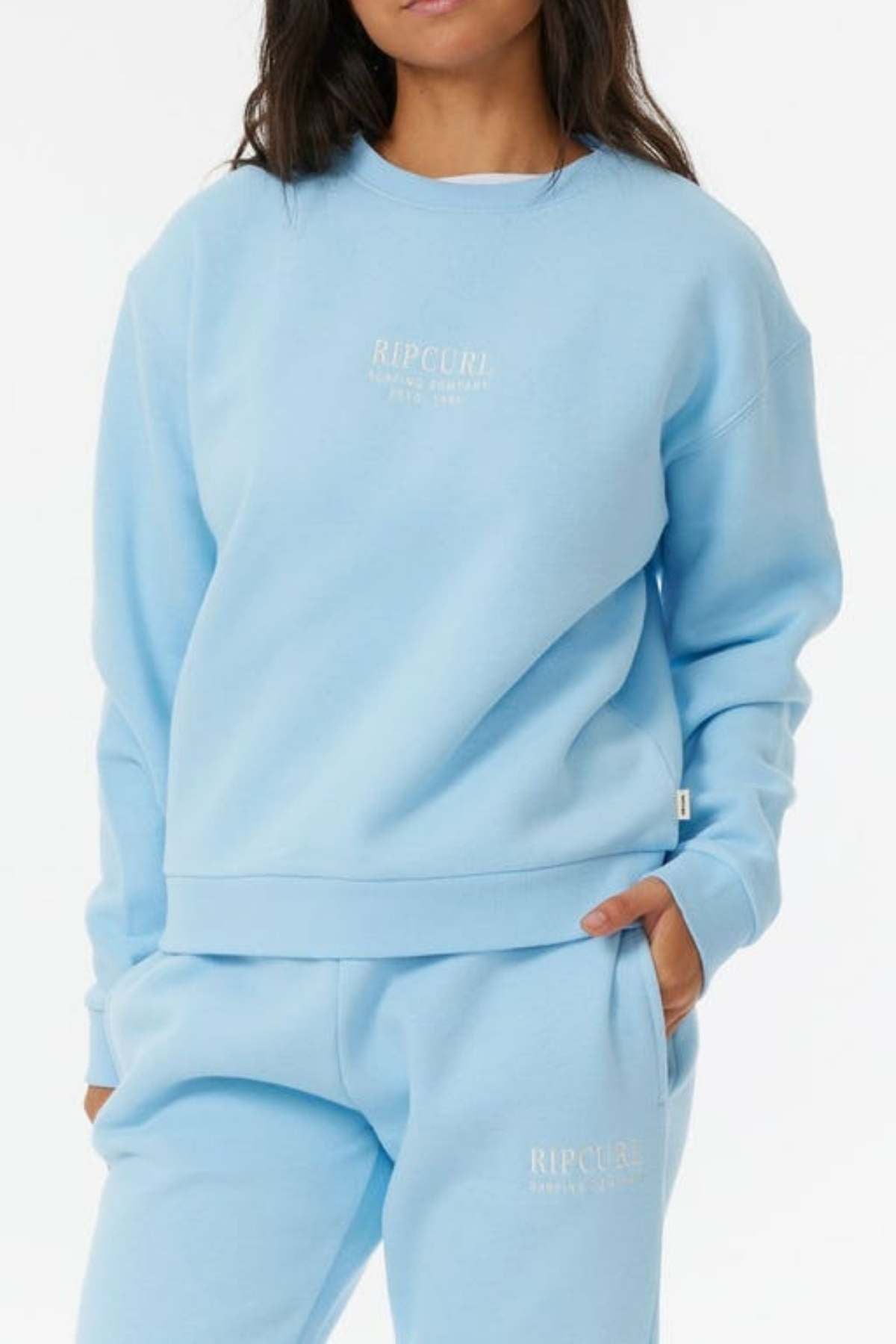 A person wearing a light blue Rip Curl Surf Staple Relaxed Crew with fleece lining and matching sweatpants, hands in pockets, stands against a white background. Both feature small white logos.