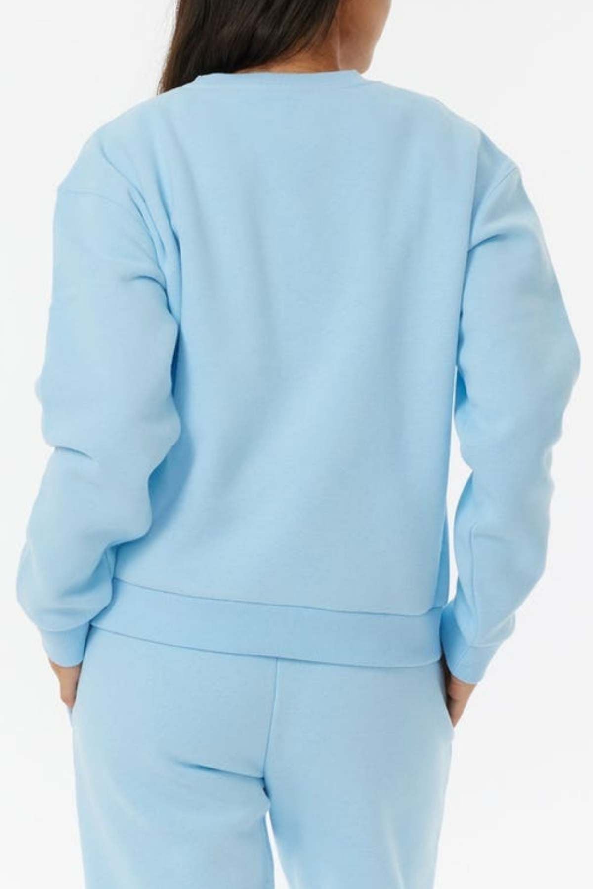 A person with long dark hair, seen from behind, wears the Rip Curl Surf Staple Relaxed Crew in light blue with matching fleece-lined sweatpants against a plain white background.