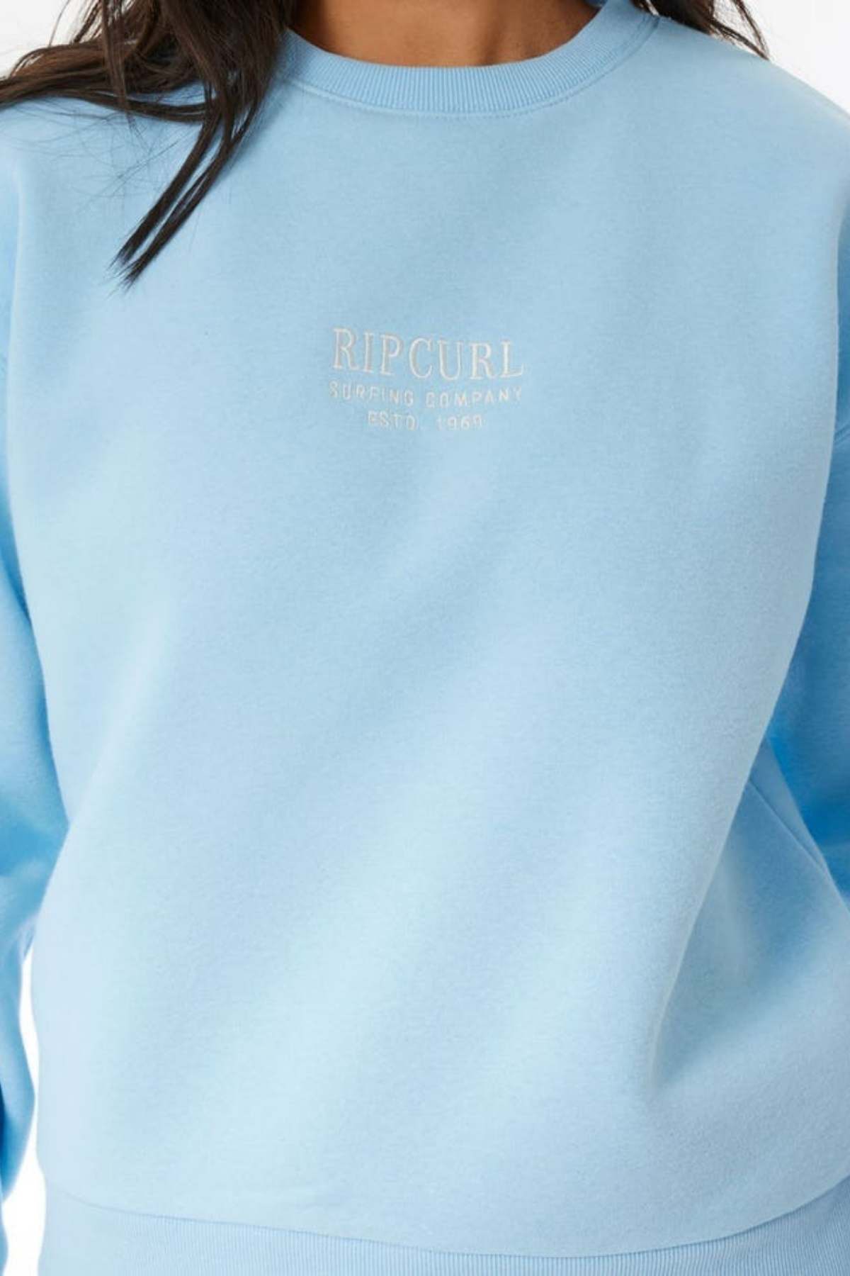 A person wears a light blue Rip Curl Surf Staple Relaxed Crew sweatshirt with the "Rip Curl Surfing Company Estd. 1969" logo in white on the chest; only their upper torso and part of their hair are visible.