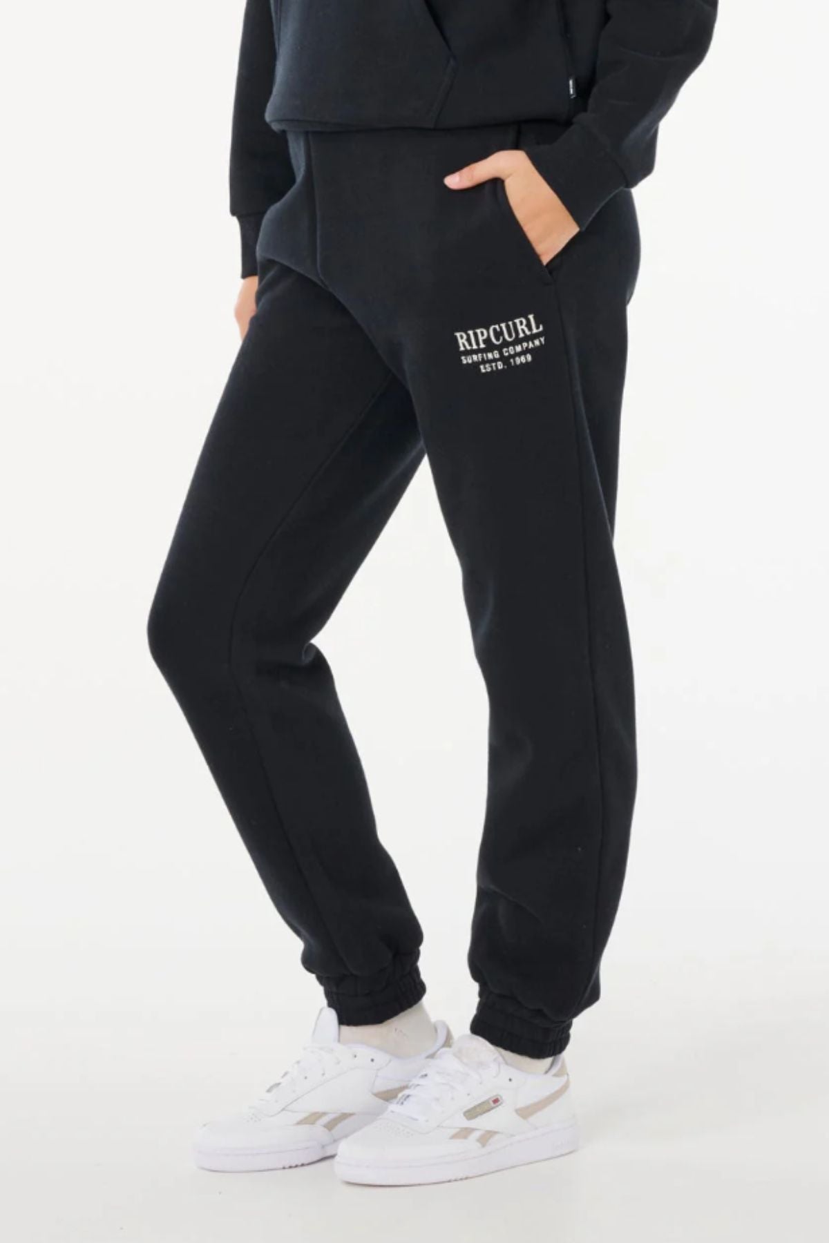 Someone stands against a plain white background wearing black Rip Curl Surf Staple Trackpants with an elastic waist, hands in pockets, paired with a black sweatshirt and white sneakers.