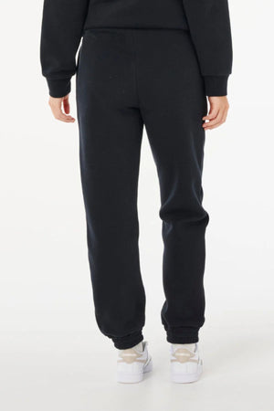 A person is standing with their back to the camera, wearing black Rip Curl Surf Staple Trackpant, a matching black sweatshirt, and white sneakers against a plain white background.
