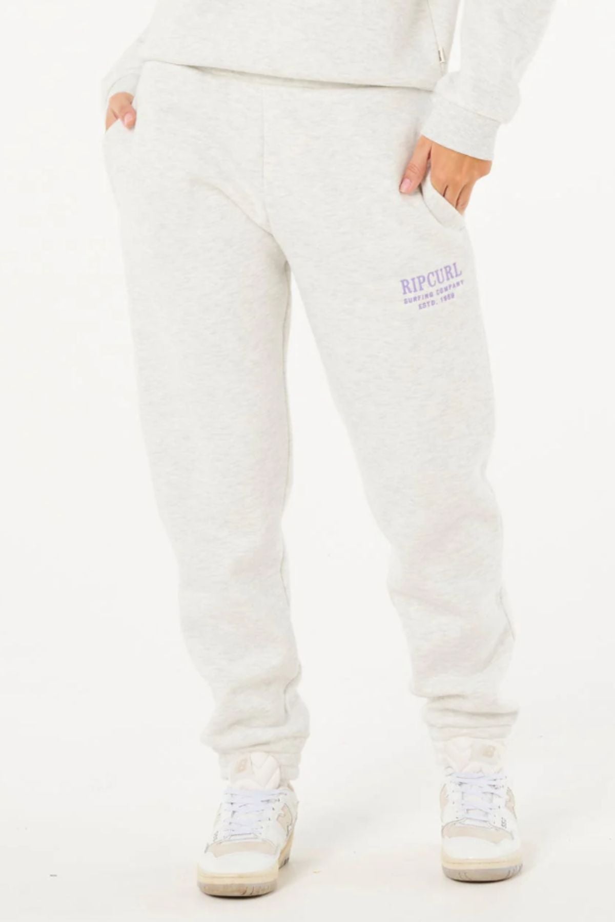 Wearing Rip Curl Surf Staple Trackpant in light gray with purple "Rip Curl" text on the left thigh, hands in pockets, styled with white sneakers—ideal casual wear against a plain white background.