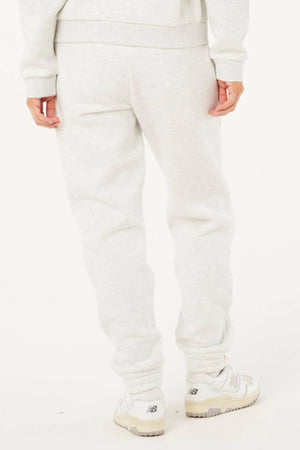 Person wearing light gray Rip Curl Surf Staple Trackpant and white sneakers, standing with their back to the camera against a plain white background. Only the lower body is visible, emphasizing relaxed, casual style.