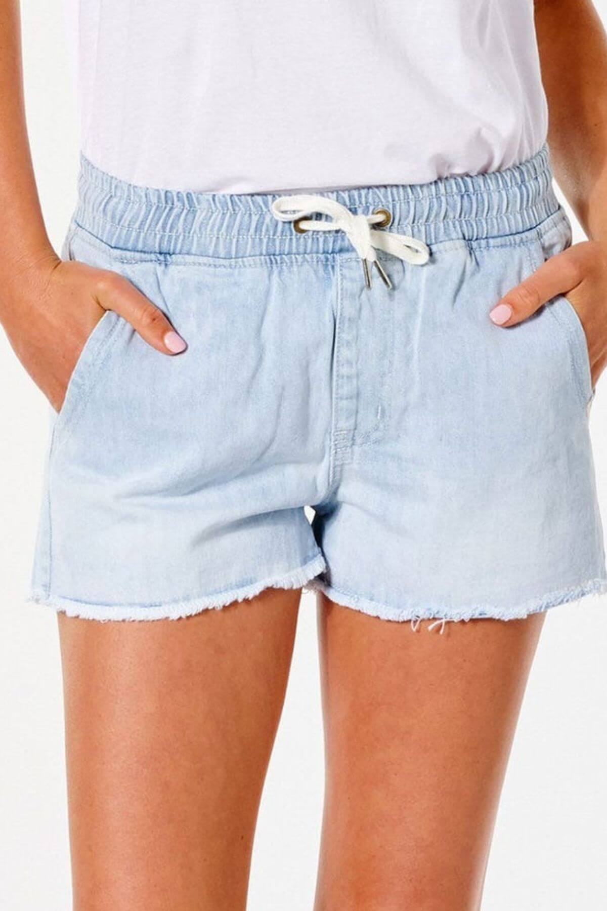 Wearing Rip Curl Tara Short vintage-inspired light blue denim shorts with a white drawstring and frayed hems, paired with a white shirt and hands in pockets—a look that's perfect for a relaxed summer day.