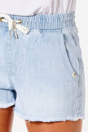 A person is wearing Rip Curl Tara Shorts featuring light-wash denim with a vintage distressed look, an elastic waistband, frayed hems, and a white drawstring. They pair it with a tucked-in white top for effortless style.