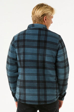 A person with blonde hair, facing away from the camera, wears a blue and black Rip Curl Tofino Polar Fleece Jacket made from recycled poly fleece, paired with black pants against a plain light background.