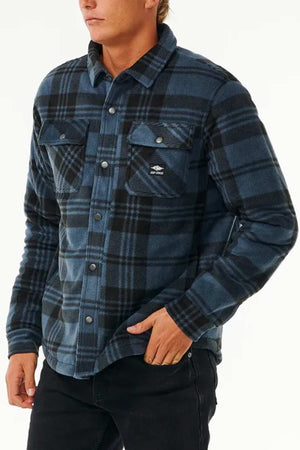 A person in a blue and black plaid button-up shirt with two chest pockets and black jeans stands against a white background. With their left hand in their pocket, they have a Rip Curl Tofino Polar Fleece Jacket draped over one shoulder for an added touch of effortless style.