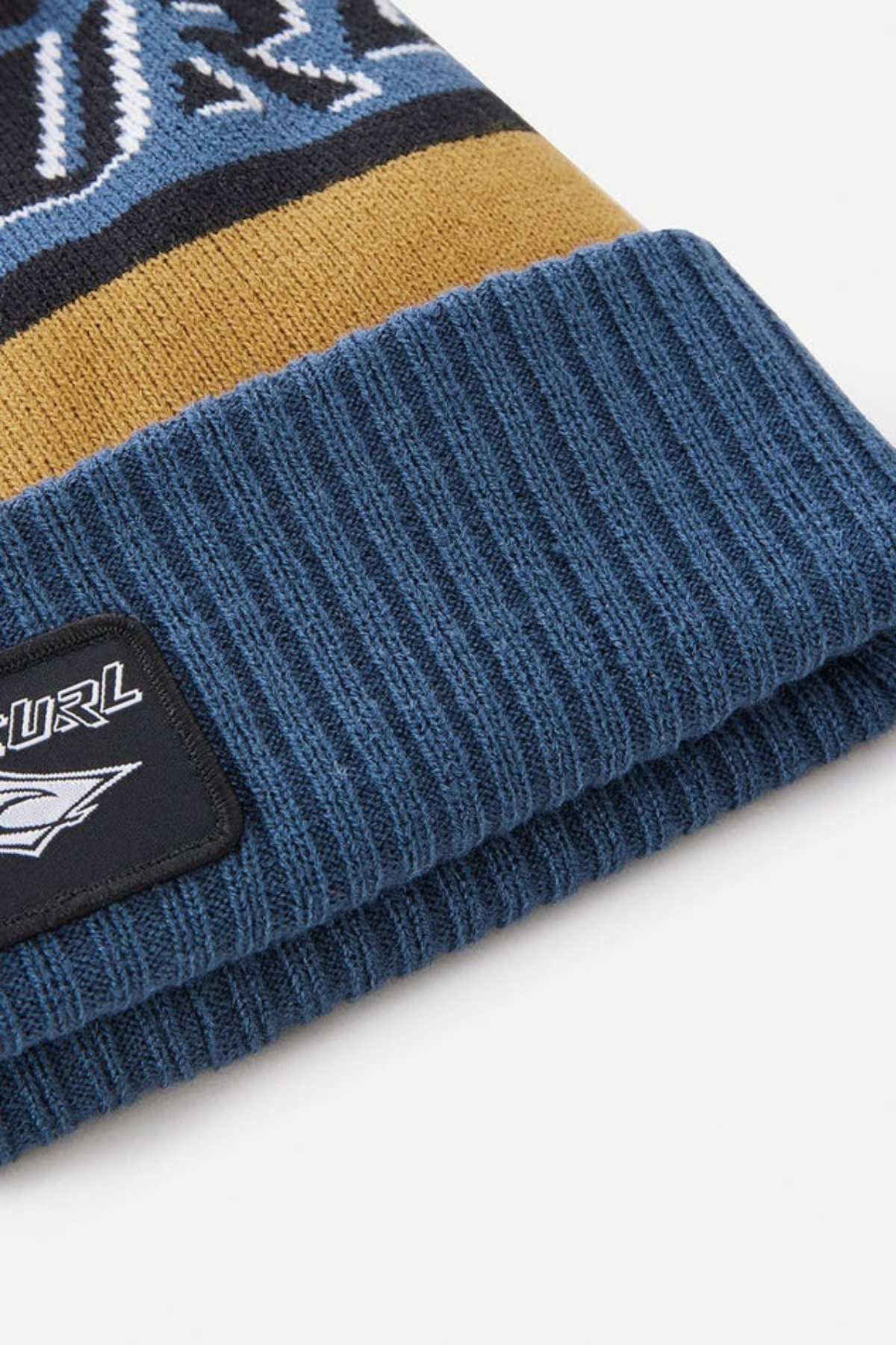 The Rip Curl Tow In Tall Beanie is a blue ribbed knit beanie made of soft acrylic yarn, featuring a yellow stripe, a black patch with the Rip Curl logo and wave design, and partial graphics in white, blue, and black at the top.