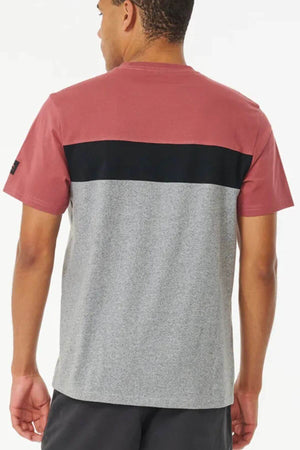 A man stands with his back to the camera, wearing a Rip Curl Undertow Panel Tee featuring mauve sleeves and upper back, a black horizontal stripe, and a heather gray lower section.