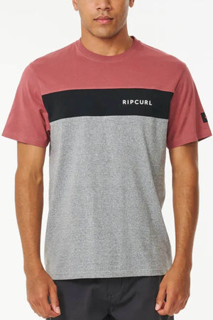 A man stands with arms relaxed, wearing the Rip Curl Undertow Panel Tee—a short-sleeve, color-blocked shirt featuring a maroon top, black stripe, light gray bottom, and a logo on the chest.