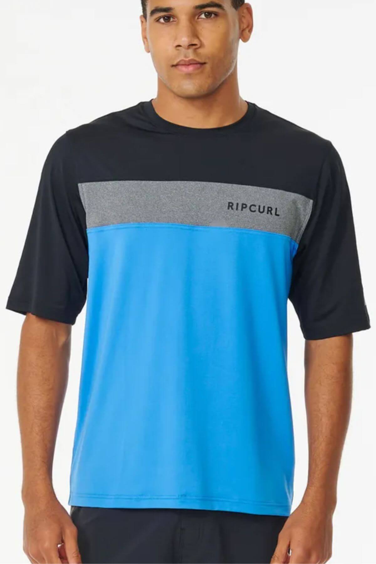 A man wearing a Rip Curl Undertow Short Sleeve UV Tee (UPF50+), with black shoulders, a gray chest stripe displaying "Rip Curl," and a bright blue lower section, stands against a plain white background.