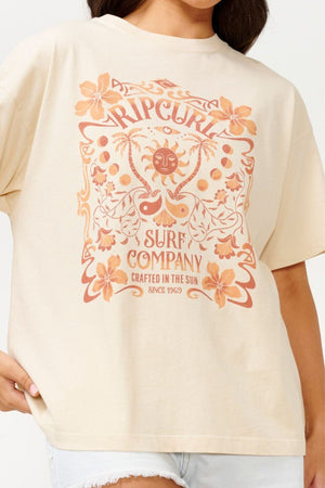 A person embraces a vintage vibe with the Rip Curl Utopian Dreams Heritage Tee, a beige cotton shirt featuring floral and sun motifs alongside the text "Rip Curl Surf Company, Crafted in the Sun, Since 1969." This short-sleeved tee, enriched with orange and brown tones, is fashionably matched with light denim shorts.