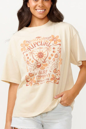 A person smiles in a Rip Curl Utopian Dreams Heritage Tee, featuring an artistic design with flowers and a sun emblem. The shirt reads "Rip Curl Surf Company Crafted by the Sun," exuding vintage charm. With long hair and light denim shorts, their relaxed style is effortlessly cool.