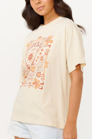 A person wearing the Rip Curl Utopian Dreams Heritage Tee, showcasing vintage charm with its orange floral and sun design on a beige background, stands against a plain backdrop. The cotton tee is effortlessly paired with light denim shorts.