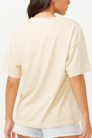 A person wearing a Rip Curl Utopian Dreams Heritage Tee in beige and light denim shorts is shown from the back against a neutral background.