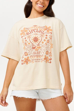 A person wearing a Rip Curl Utopian Dreams Heritage Tee, which features a vintage charm in beige with an orange floral design and the text "Surf Company," is smiling. They are also wearing white denim shorts.