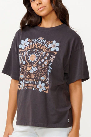 A person wearing the Rip Curl Utopian Dreams Heritage Tee, featuring a dark base with a floral and sun design and the text "Rip Curl Surf Company," exudes vintage charm. This heritage fit pairs perfectly with light-colored pants, as the individual with long dark hair stands against a plain background.