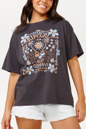 A person wearing the Rip Curl Utopian Dreams Heritage Tee, which features an artistic graphic design with "RIP CURL Surf Company," floral patterns, and a sun motif in a dark hue, evokes vintage charm. Paired with light denim shorts, this outfit perfectly captures the essence of Rip Curl Utopian Dreams.