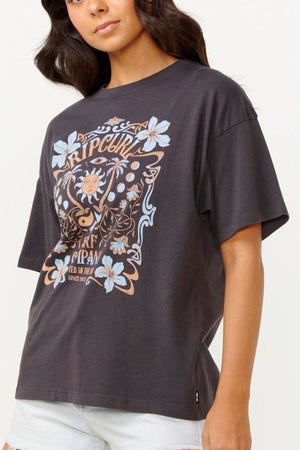 A woman in a loose-fitting black Rip Curl Utopian Dreams Heritage Tee, featuring floral elements and vintage charm, pairs it with light-colored shorts against a plain, light background.