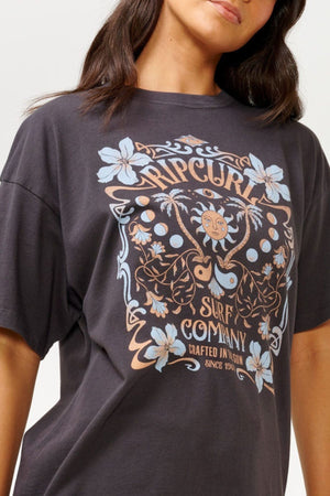 A woman wears the Rip Curl Utopian Dreams Heritage Tee, which exudes vintage charm with its colorful floral designs and text. The shirt's earthy tones and decorative elements perfectly capture the Rip Curl Utopian Dreams vibe. Her dark, wavy hair beautifully complements the heritage fit.