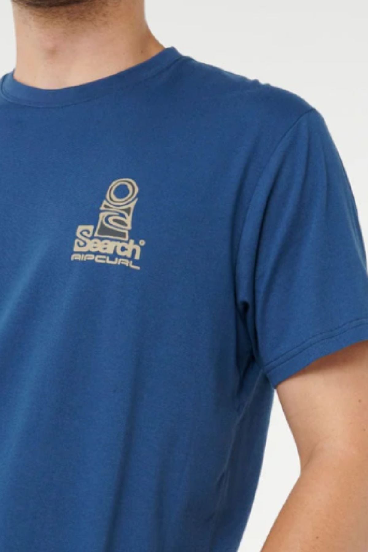 A person wearing the Rip Curl Vaporcool Search Stack Tee—a blue moisture-wicking shirt featuring a small beige "Search" logo on the chest. The image shows their upper torso and part of the right arm.