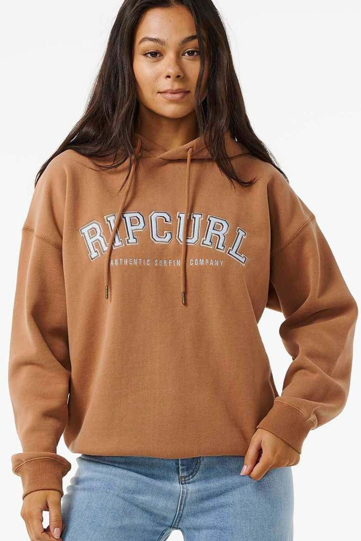 A person with long dark hair wears an oversized Rip Curl Varsity Hoodie, featuring "Ripcurl Authentic Surfing Company" and a soft fleece lining, paired with blue jeans, standing against a plain background.