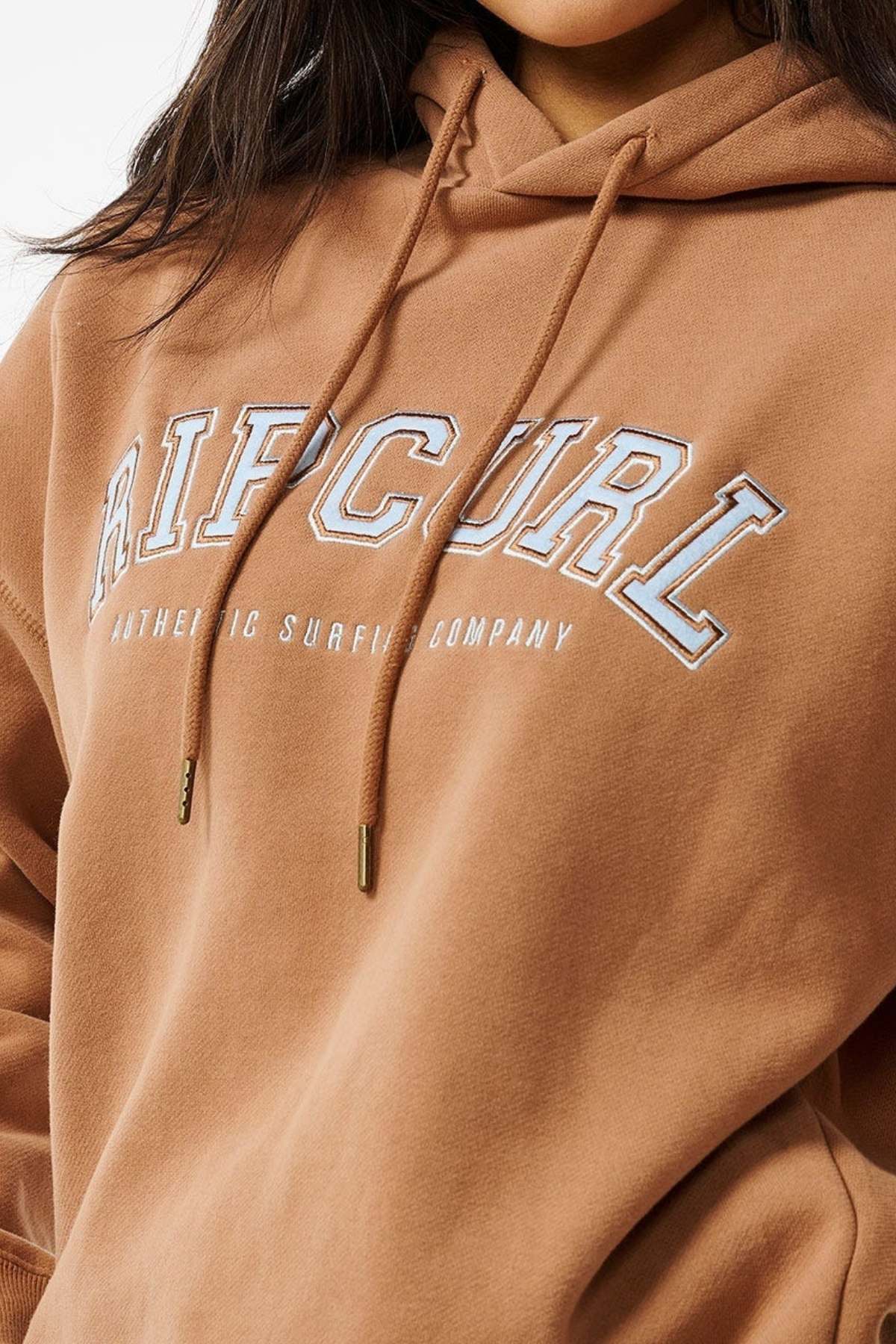 Someone is wearing a Rip Curl Varsity Hoodie in brown, featuring "Authentic Surfing Company" printed in white on the front. It has visible drawstrings and an oversized fit with a soft fleece lining, all against a plain white background.