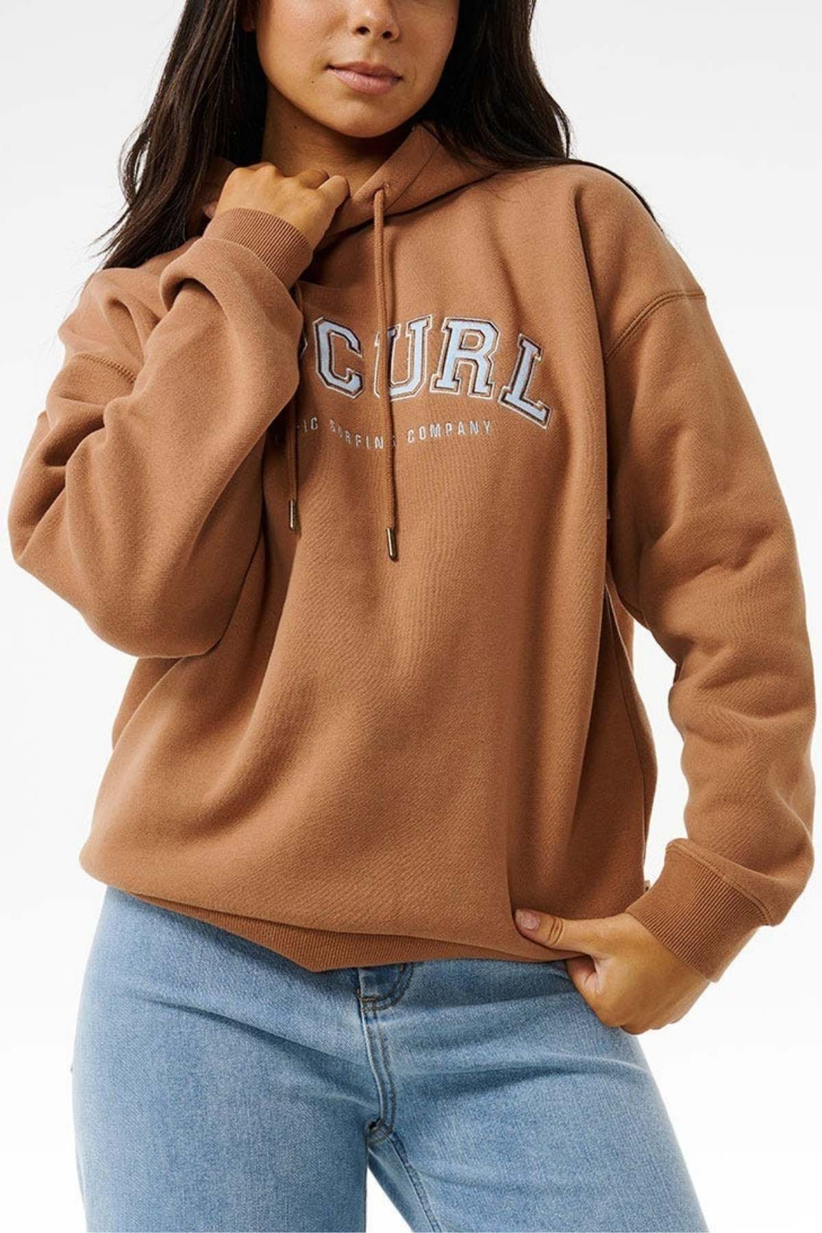 A person in a brown Rip Curl Varsity Hoodie with "CURL" on it and light blue jeans stands relaxed against a plain background. The hoodie, with an adjustable drawstring hood, offers an oversized fit and soft fleece lining for extra comfort.