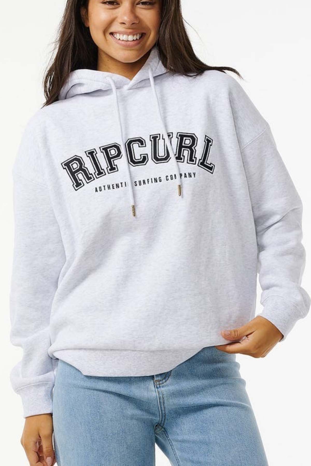 A woman smiles in the Rip Curl Varsity Hoodie, featuring an oversized fit, light gray fleece fabric, bold front text, and a front pocket. She pairs it with blue jeans and stands against a plain white background.