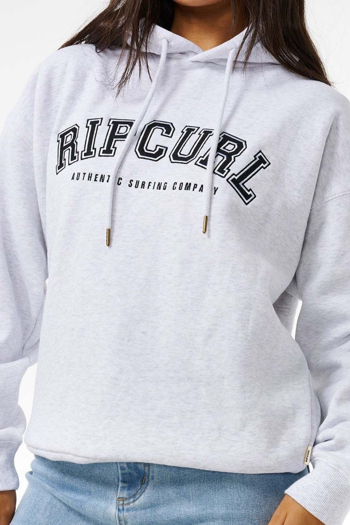 A person wears a light grey Rip Curl Varsity Hoodie with "AUTHENTIC SURFING COMPANY" text, featuring an oversized fit, paired with light blue jeans. Their face is partially out of frame.