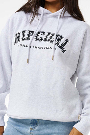 A person wears a light grey Rip Curl Varsity Hoodie with "AUTHENTIC SURFING COMPANY" text, featuring an oversized fit, paired with light blue jeans. Their face is partially out of frame.