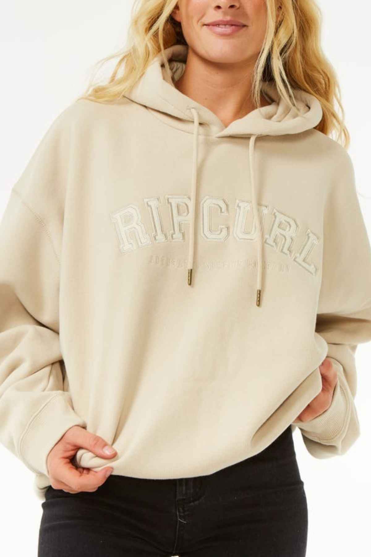 Wearing a beige Rip Curl Varsity Hoodie with the hood up and hands gripping the Cotton Poly Fleece hem, this person pairs it with black pants. Against a white background, the relaxed Heritage fit stands out.