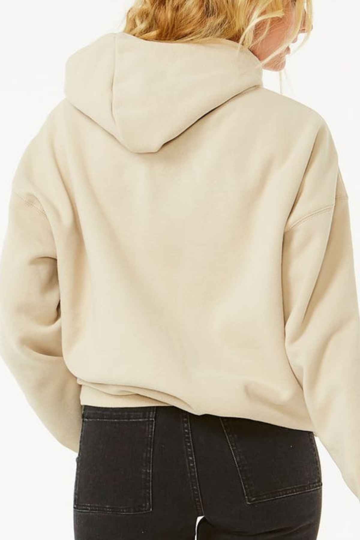 A person with long, wavy blond hair is wearing a Rip Curl Varsity Hoodie in light beige Heritage fit and black jeans, facing away from the camera against a plain white background.