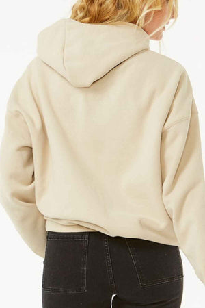A person with long, wavy blond hair is wearing a Rip Curl Varsity Hoodie in light beige Heritage fit and black jeans, facing away from the camera against a plain white background.