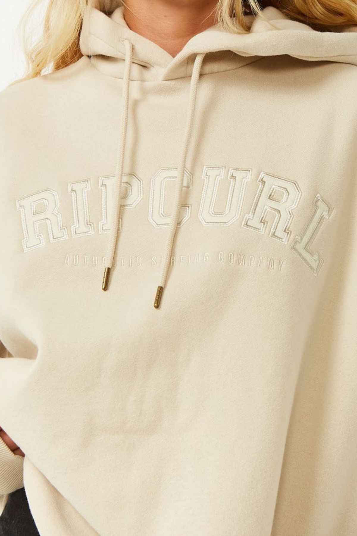 A person wears the Rip Curl Varsity Hoodie, heritage fit, made from Cotton Poly Fleece, with "RIP CURL" and "AUTHENTIC SURFING COMPANY" embroidered in white on the front. Only their lower face, torso, and part of hair are visible.