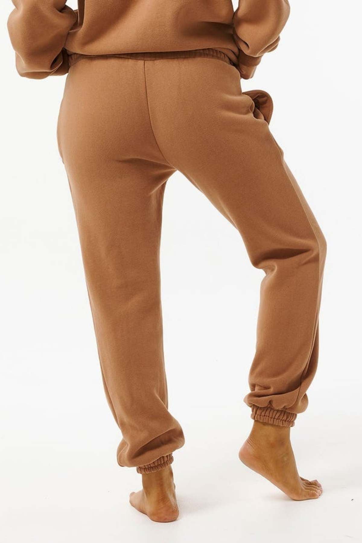 Wearing Rip Curl Varsity Pants made from a soft cotton/polyester blend, a barefoot person stands on a light surface with one leg slightly bent and hands in pockets, highlighting the pants’ comfort and casual style against a plain white background.