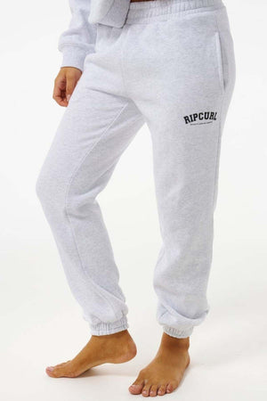 Wearing Rip Curl Varsity Pants in light gray with elastic cuffs, a black Rip Curl logo on the left hip, and made from soft Cotton Poly Fleece; model is barefoot against a white background.