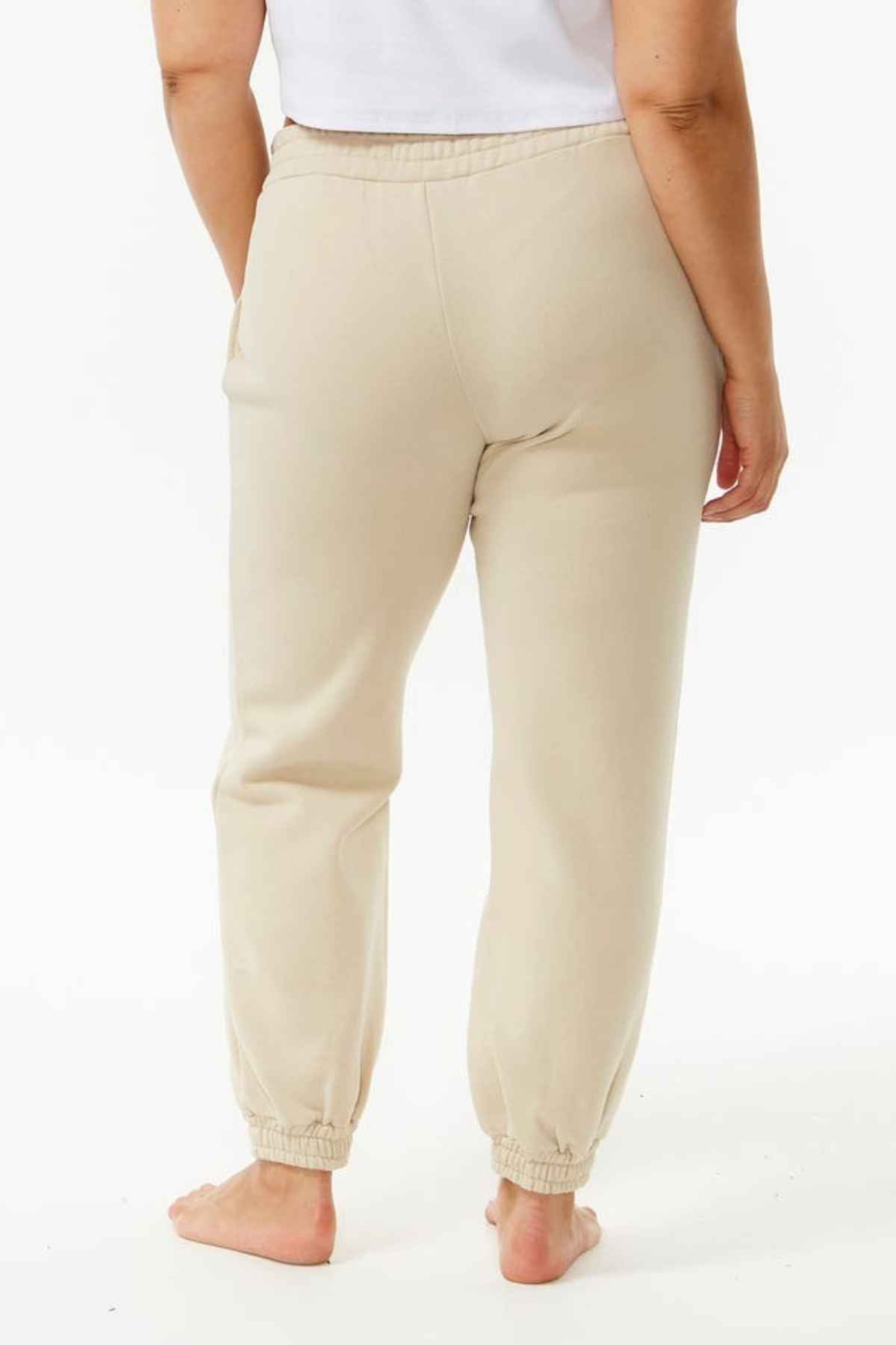 A person stands barefoot facing away, wearing Rip Curl Varsity Pants—a beige jogger-style sweatpant made from soft cotton poly fleece—with a white top, set against a plain white background to highlight this casual outfit.