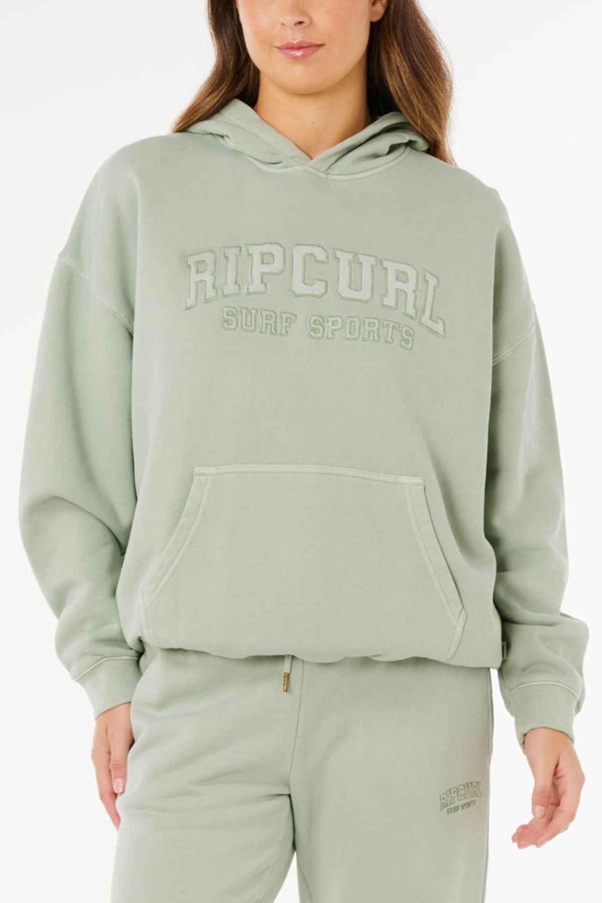 A woman models the Rip Curl Varsity Sports Oversized Hood in light green, made from 100% cotton with a front pocket and embroidered chest text, paired with matching sweatpants against a plain white background.
