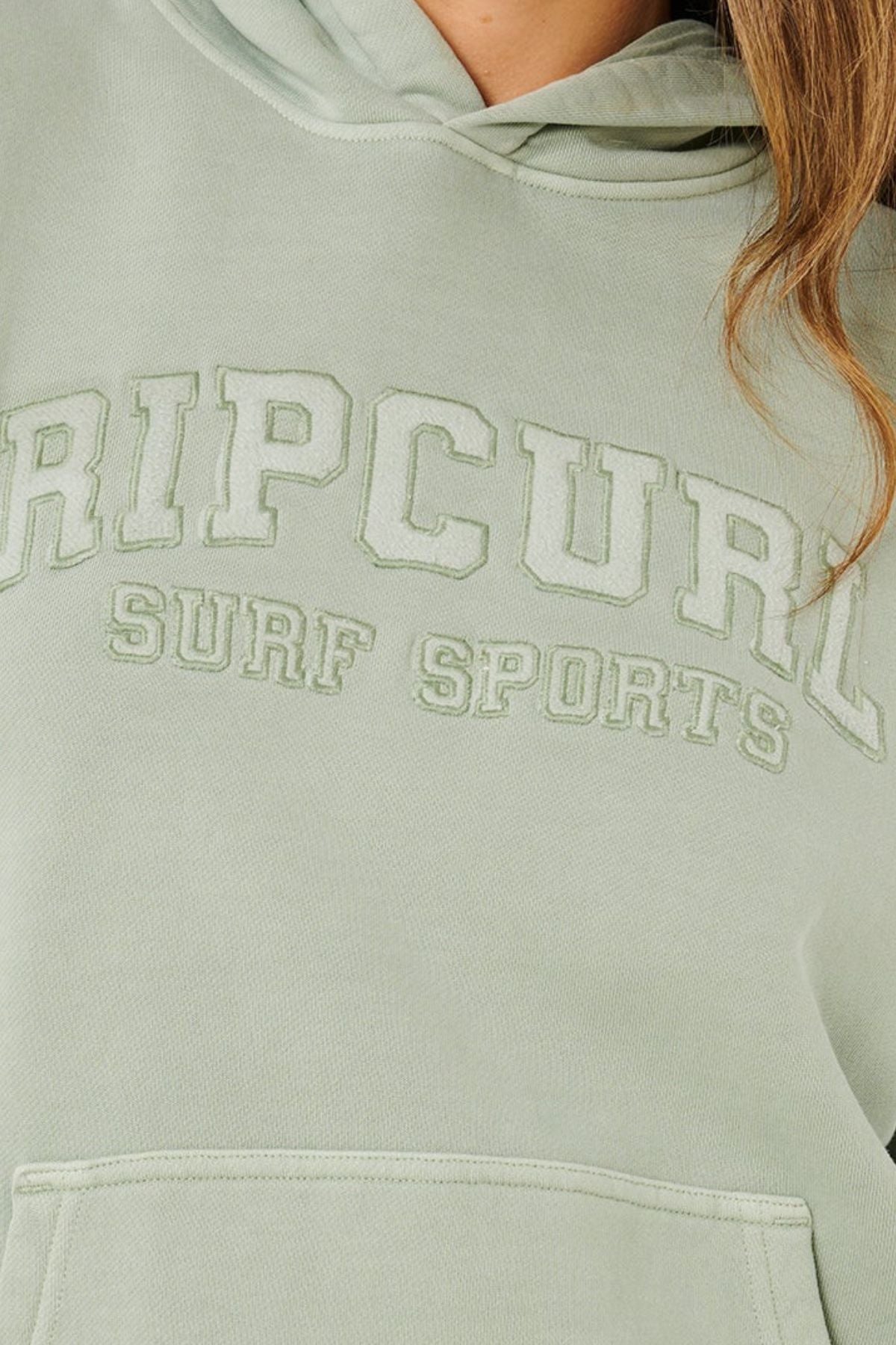 A person in a light green Rip Curl Varsity Sports Oversized Hood, featuring "RIP CURL SURF SPORTS" embroidery on the front. Only part of their face and hair visible. Made from 100% cotton for comfort.
