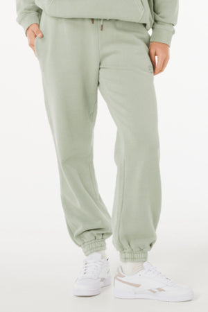 Wearing light green Rip Curl Varsity Sports Track Pant, a matching sweatshirt, and white sneakers, a person stands with hands in pockets against a plain white background.
