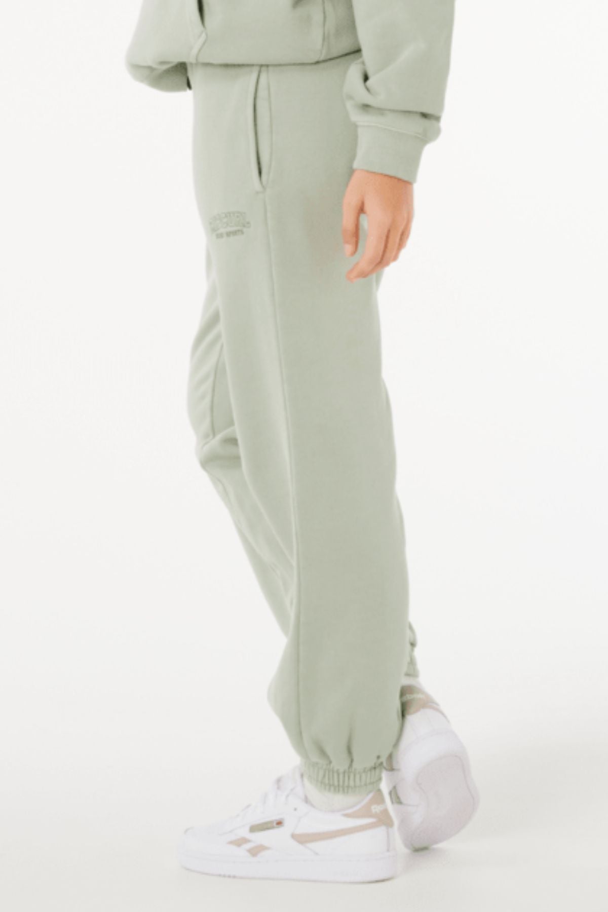 Someone stands sideways against a plain white background, wearing light green Rip Curl Varsity Sports Track Pant made from 100% cotton and white sneakers, with one hand by their side.