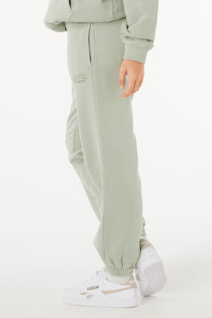 Someone stands sideways against a plain white background, wearing light green Rip Curl Varsity Sports Track Pant made from 100% cotton and white sneakers, with one hand by their side.