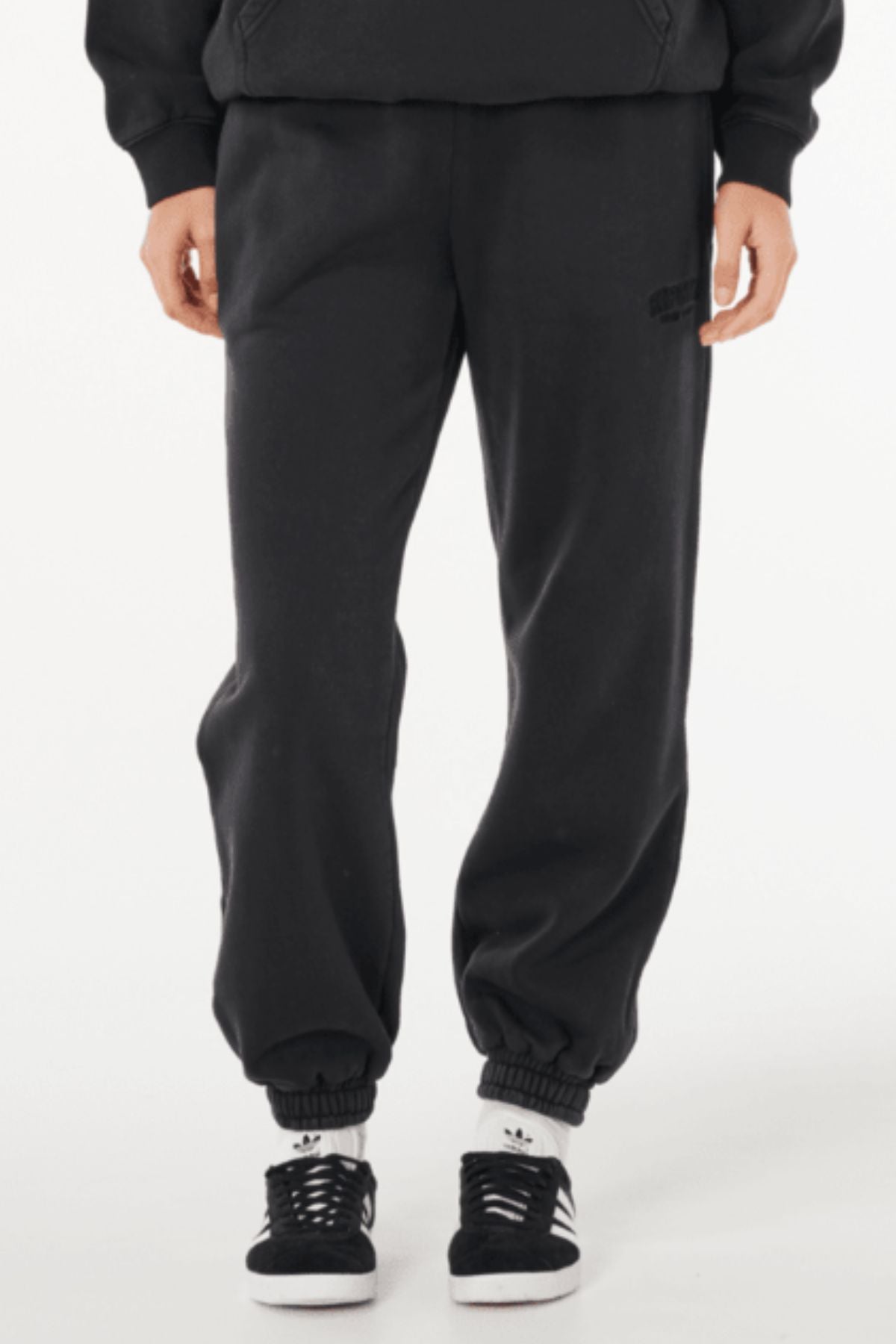 A person wearing Rip Curl Varsity Sports Track Pant in black, a black sweatshirt, and black-and-white Adidas sneakers stands against a plain white background.