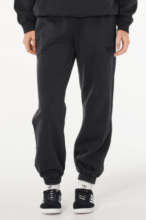 A person wearing Rip Curl Varsity Sports Track Pant in black, a black sweatshirt, and black-and-white Adidas sneakers stands against a plain white background.